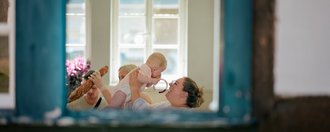MuKi Haus Alfter Eine Frau hält ein Baby vor sich hoch. Sie sitzt an einem Tisch in der Küche. Es wurde von draußen durch das offene Fenster fotografiert.