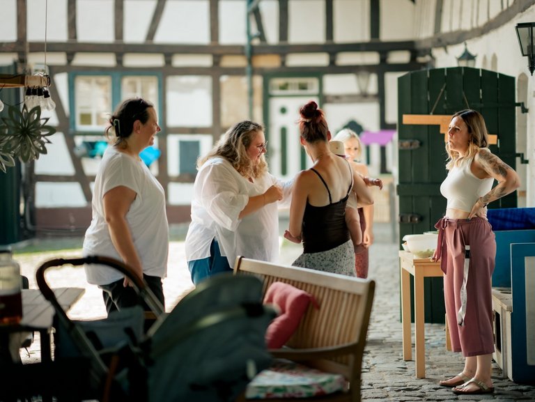 Eine Gruppe Frauen mit einem Baby steht im Innenhof eines Bauernhofes und unterhalten sich.