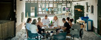 MuKi Haus Alfter Eine Gruppe Menschen sitzt im Hof eines Bauernhofes um einen Tisch beim Essen.